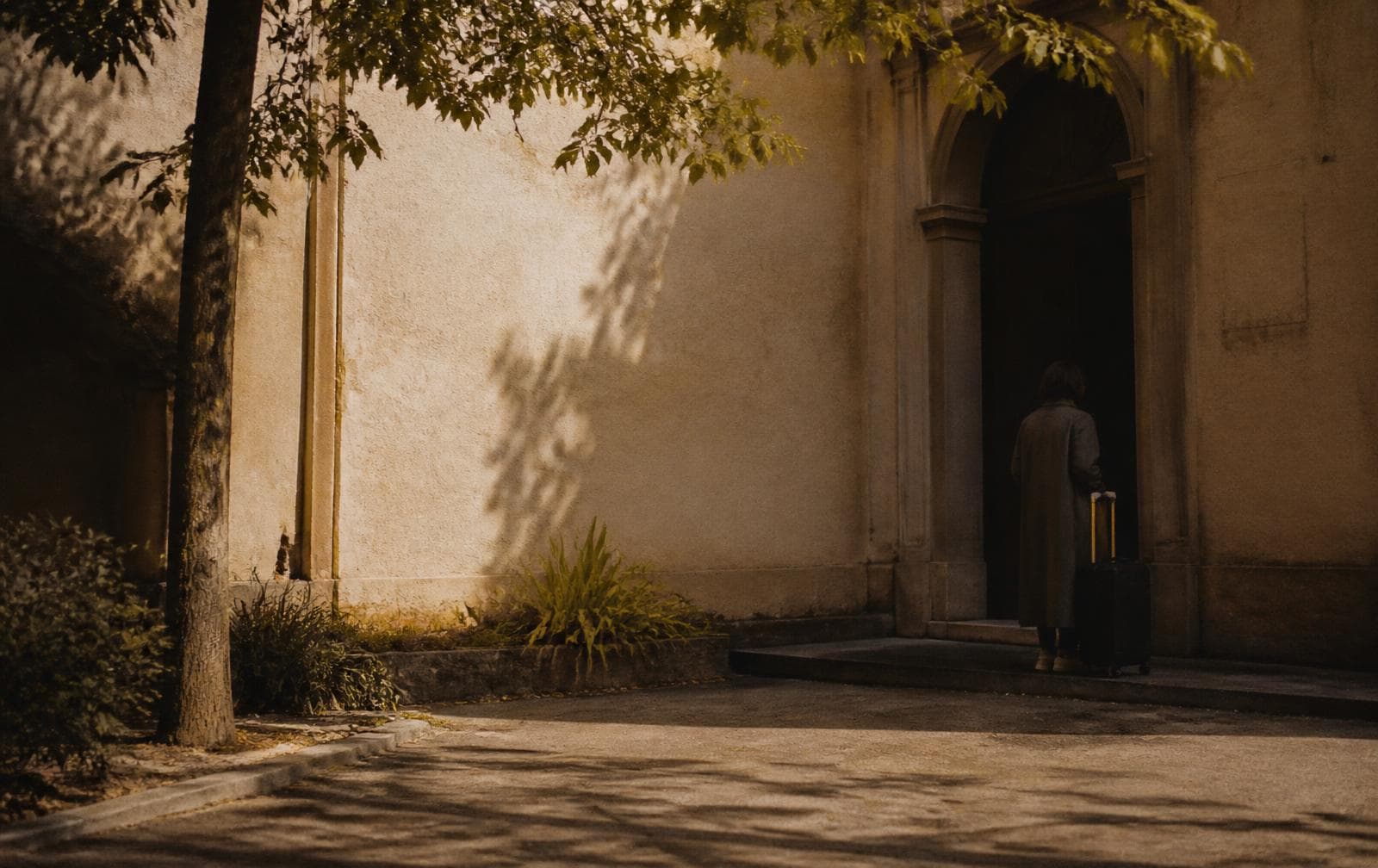 A person waiting quietly at a shaded entrance with a small suitcase