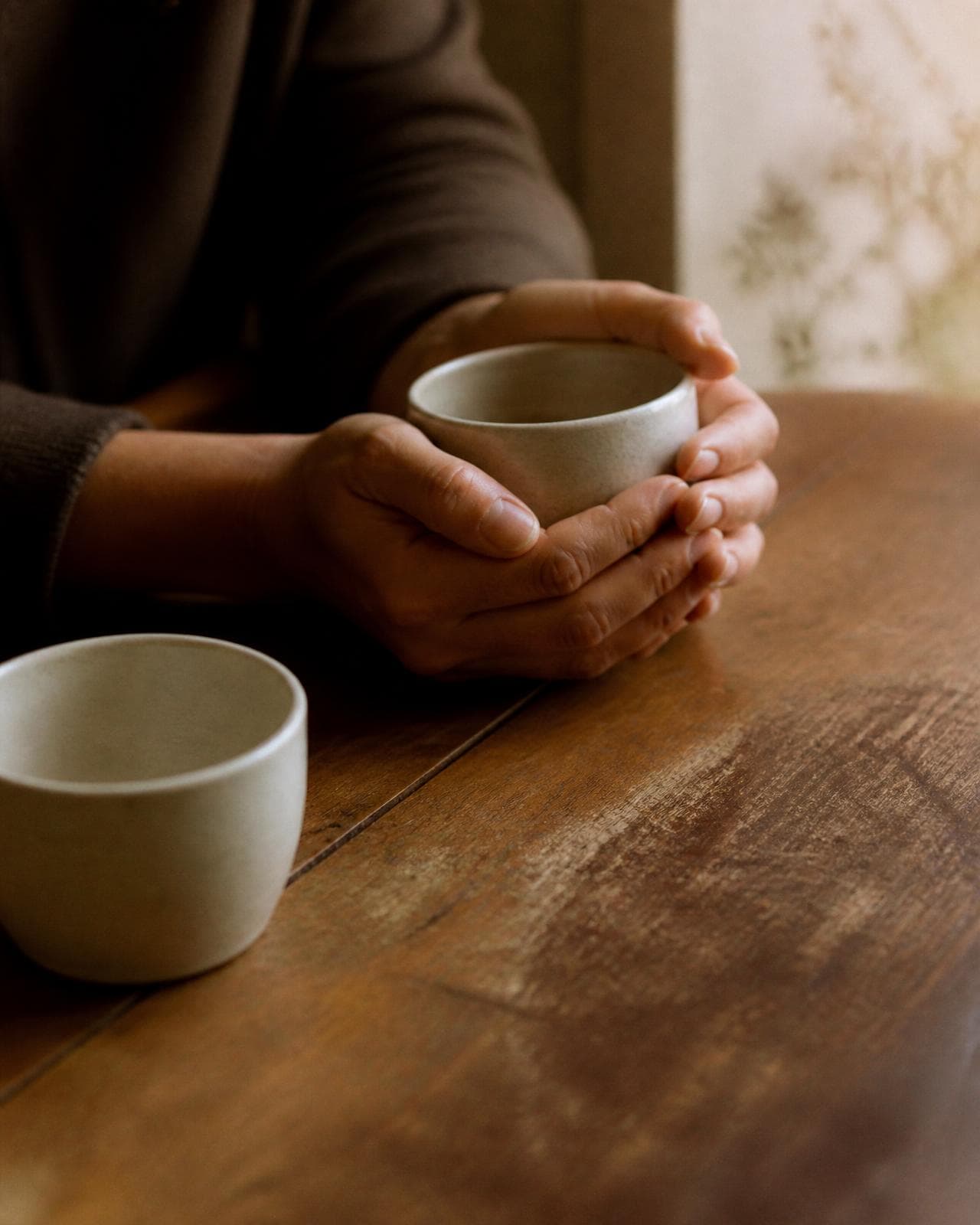 Two hands resting around a cup on a wooden table, a second cup nearby