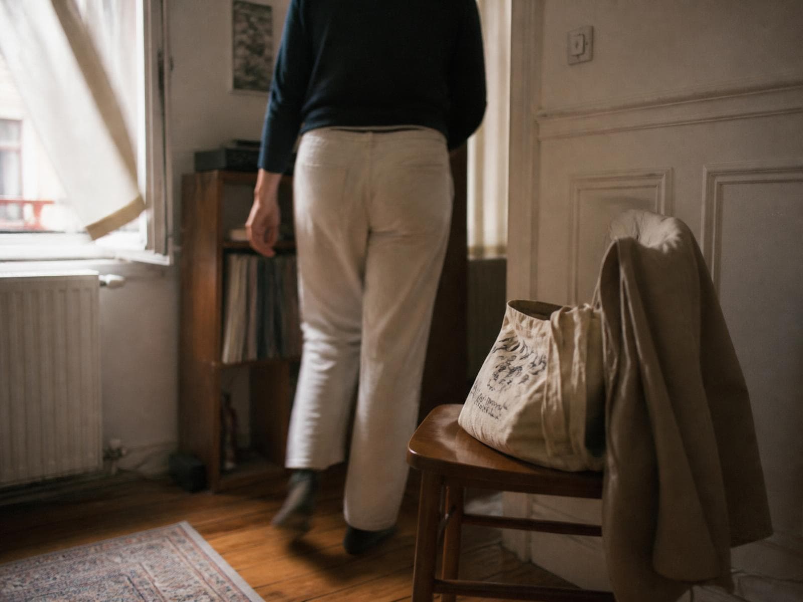 Someone walking through a lived-in room in daylight, a bag set down on a chair with a coat over its back