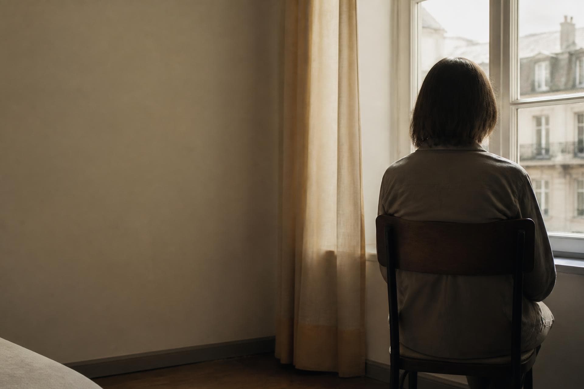 Someone sitting quietly by a window in an ordinary room, seen from behind